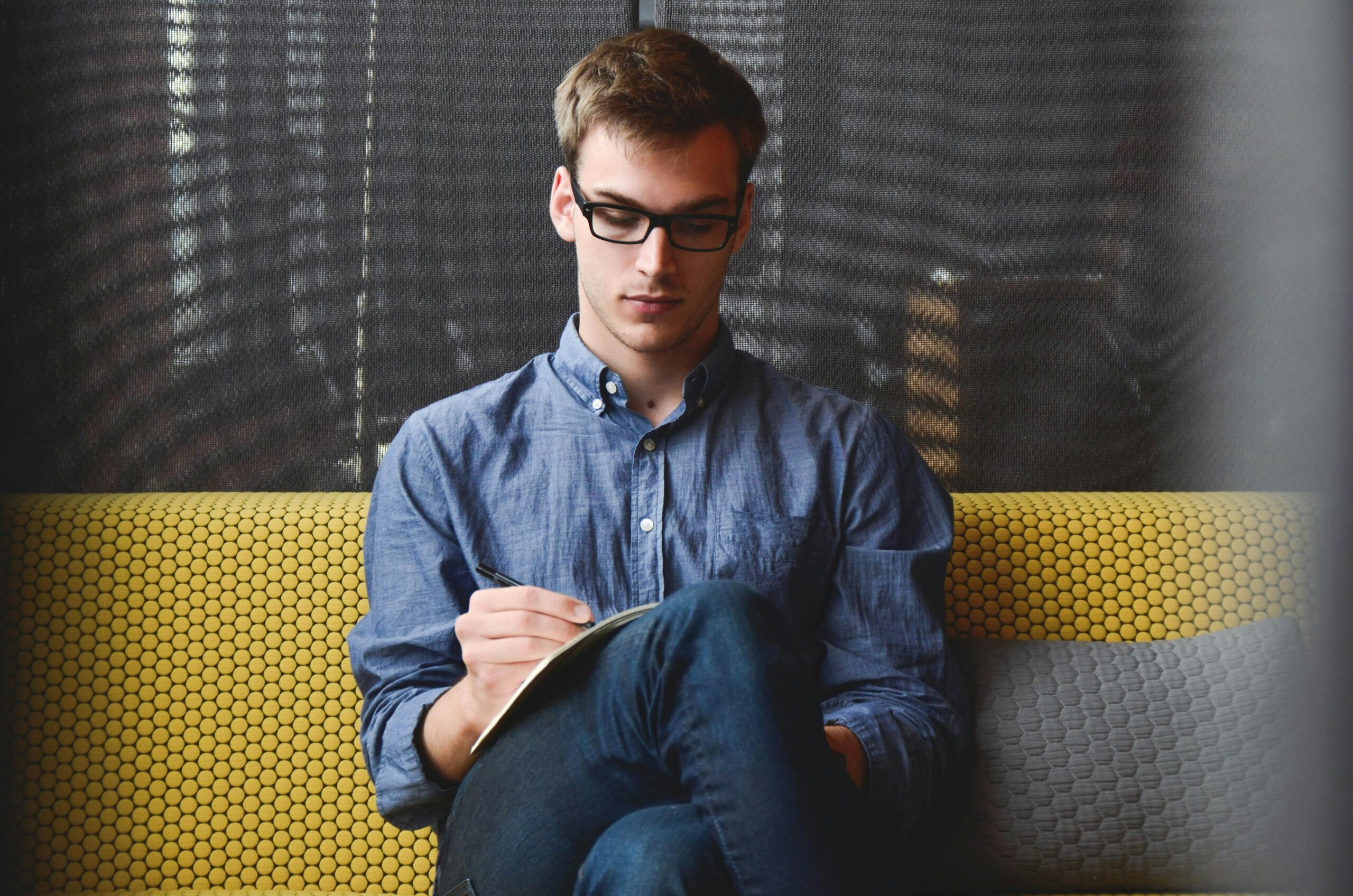 Inicio A young man in glasses writes in a notebook while sitting on a stylish couch indoors.
